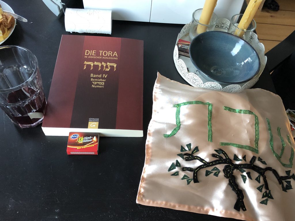 A photograph of my shabbat set up. A glass with grape juice, a plate with bread covered with a satin cloth that has the Hebrew word "Shabbat" sewed onto it along with a tree of life, two candles, matches and a German version of the Torah, Bemidbar (fourth scroll of Moses) 